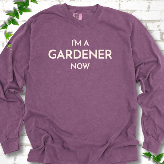 Berry long sleeve T-shirt laid flat on white brick with greenery, showing the cream words 'I'm a Gardener Now' centered on the chest.