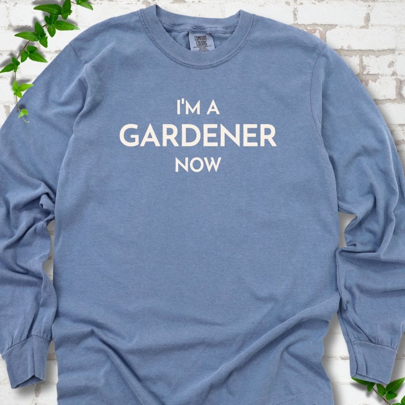 Blue Jean long sleeve T-shirt laid flat on white brick with greenery, showing the cream words 'I'm a Gardener Now' centered on the chest.