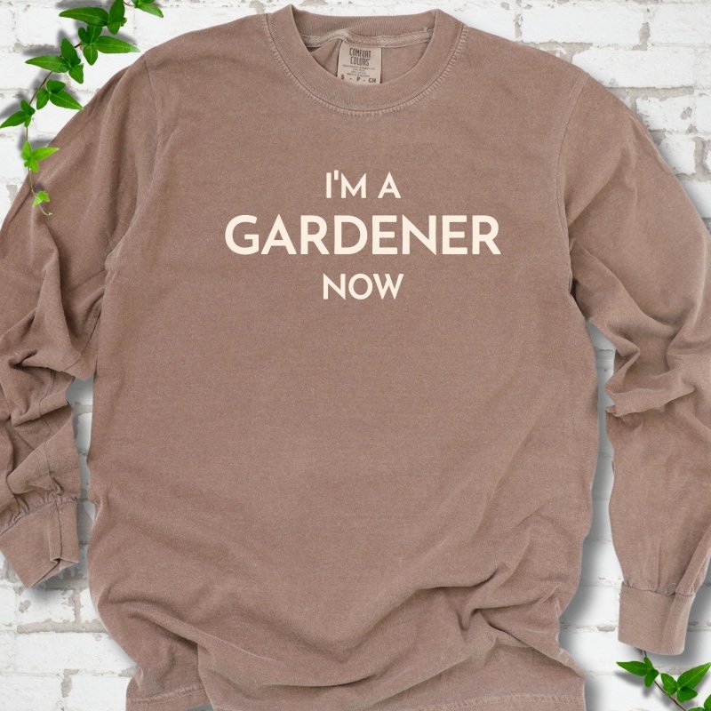 Espresso long sleeve T-shirt laid flat on white brick with greenery, showing the cream words 'I'm a Gardener Now' centered on the chest.