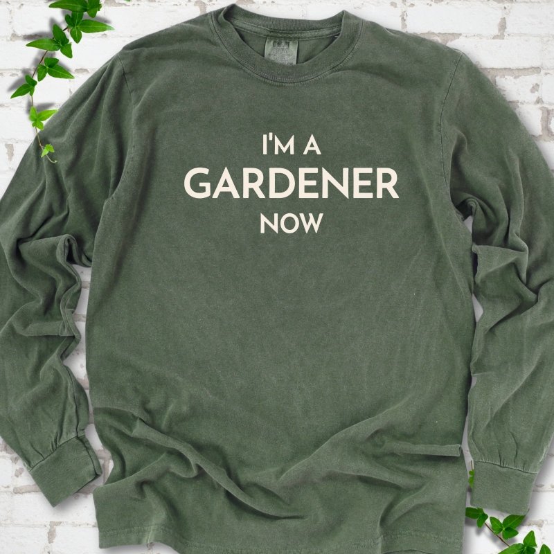 Hemp long sleeve T-shirt laid flat on white brick with greenery, showing the cream words 'I'm a Gardener Now' centered on the chest.