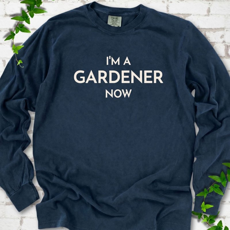 Midnight long sleeve T-shirt laid flat on white brick with greenery, showing the cream words 'I'm a Gardener Now' centered on the chest.