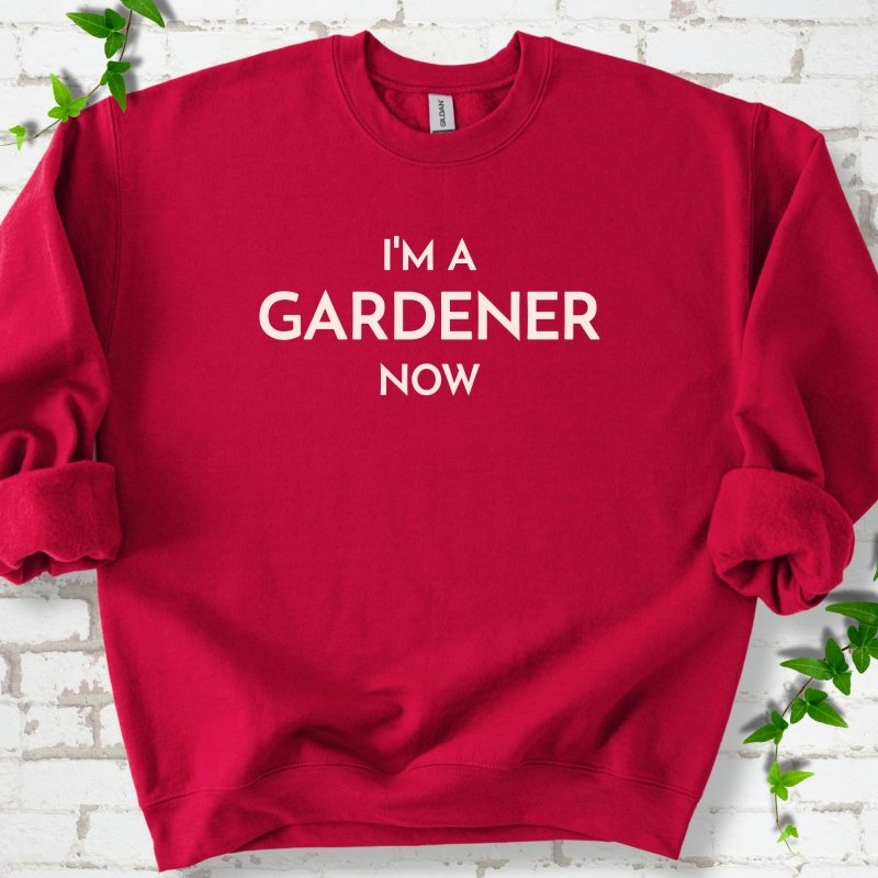 Cardinal Red crewneck sweatshirt laid flat on white brick with greenery, showing the cream words 'I'm a Gardener Now' centered on the chest.