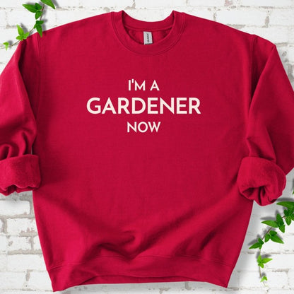 Cardinal Red crewneck sweatshirt laid flat on white brick with greenery, showing the cream words 'I'm a Gardener Now' centered on the chest.