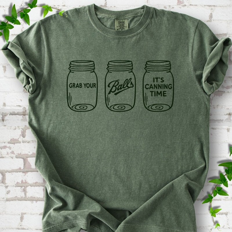 Moss T-shirt flat lay with dark green line-art illustrations of three mason jars in a row, the first reading "Grab your", the middle with "Balls" in script and the last reading "It's canning time", on white brick with ivy sprigs.
