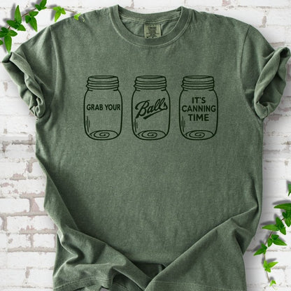 Moss T-shirt flat lay with dark green line-art illustrations of three mason jars in a row, the first reading "Grab your", the middle with "Balls" in script and the last reading "It's canning time", on white brick with ivy sprigs.