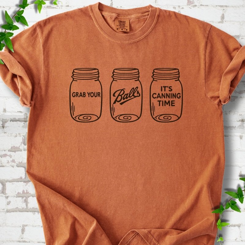 Yam T-shirt flat lay with black line-art illustrations of three mason jars in a row, the first reading "Grab your", the middle with "Balls" in script and the last reading "It's canning time", on white brick with ivy sprigs.
