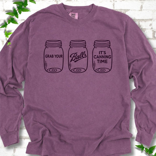 Berry long sleeve T-shirt flat lay with dark plum line-art illustrations of three mason jars in a row, the first reading "Grab your", the middle with "Balls" in script and the last reading "It's canning time", on white brick with ivy sprigs.