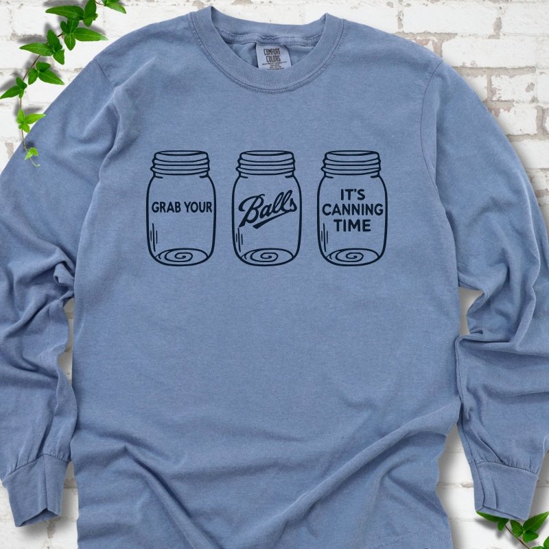 Blue Jean long sleeve T-shirt flat lay with navy line-art illustrations of three mason jars in a row, the first reading "Grab your", the middle with "Balls" in script and the last reading "It's canning time", on white brick with ivy sprigs.