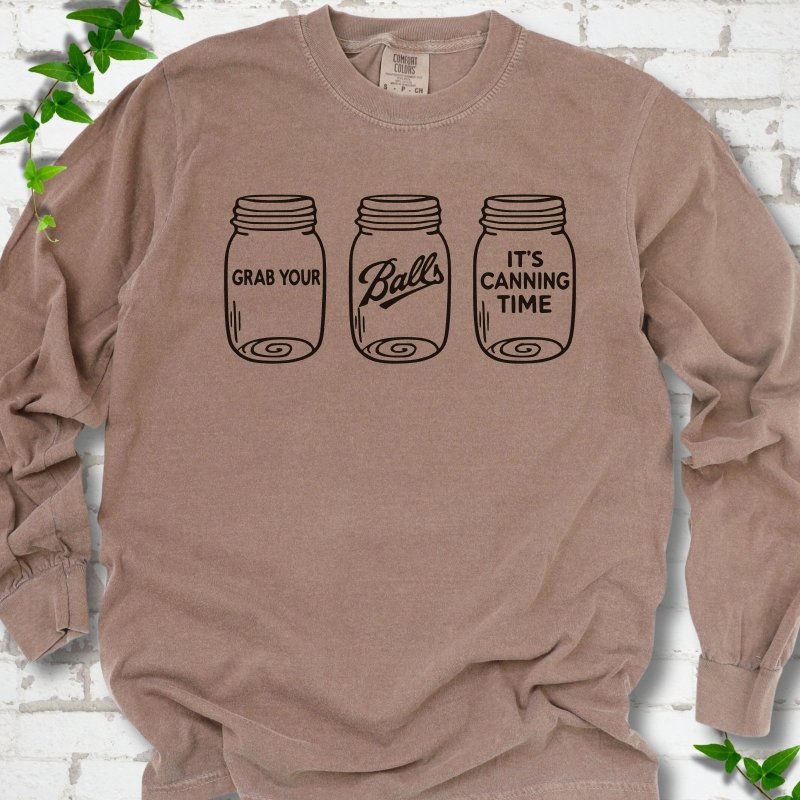 Espresso long sleeve T-shirt flat lay with black line-art illustrations of three mason jars in a row, the first reading "Grab your", the middle with "Balls" in script and the last reading "It's canning time", on white brick with ivy sprigs.