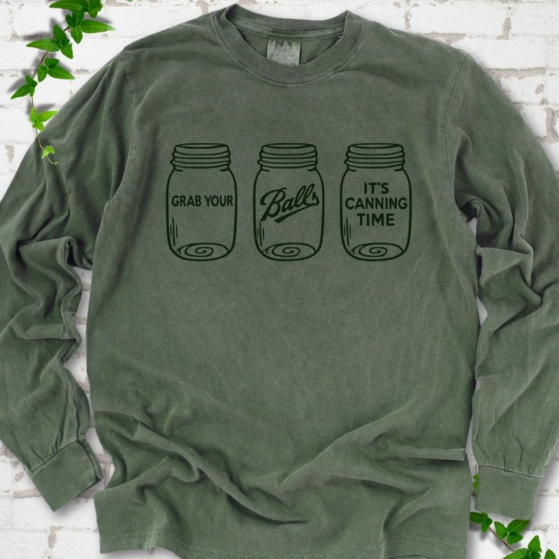 Hemp long sleeve T-shirt flat lay with dark green line-art illustrations of three mason jars in a row, the first reading "Grab your", the middle with "Balls" in script and the last reading "It's canning time", on white brick with ivy sprigs.