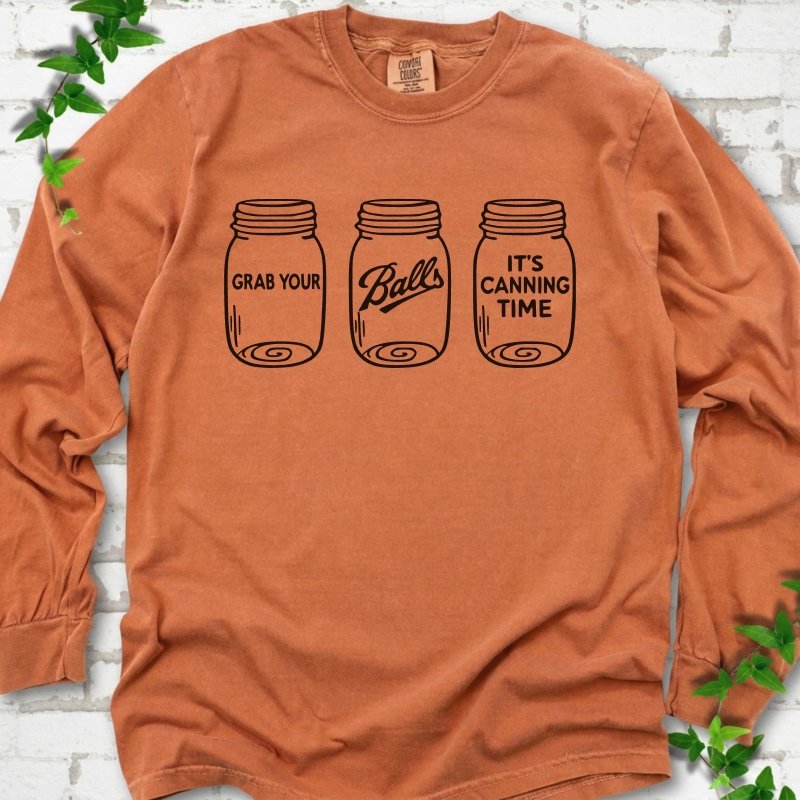 Yam long sleeve T-shirt flat lay with black line-art illustrations of three mason jars in a row, the first reading "Grab your", the middle with "Balls" in script and the last reading "It's canning time", on white brick with ivy sprigs.