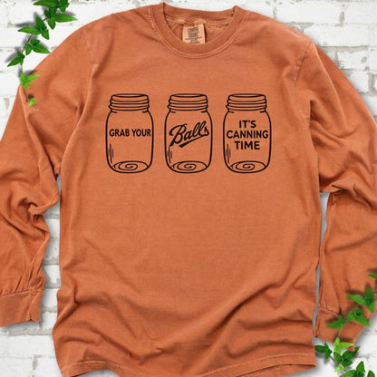 Yam long sleeve T-shirt flat lay with black line-art illustrations of three mason jars in a row, the first reading "Grab your", the middle with "Balls" in script and the last reading "It's canning time", on white brick with ivy sprigs.