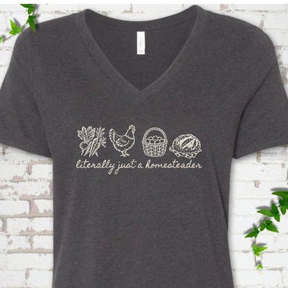 Dark Grey Heather women's V-neck T-shirt flat lay with cream line-art icons in a row of carrots and radishes, a hen, a basket of eggs and a loaf of bread above the script words "literally just a homesteader", on white brick with ivy sprigs.