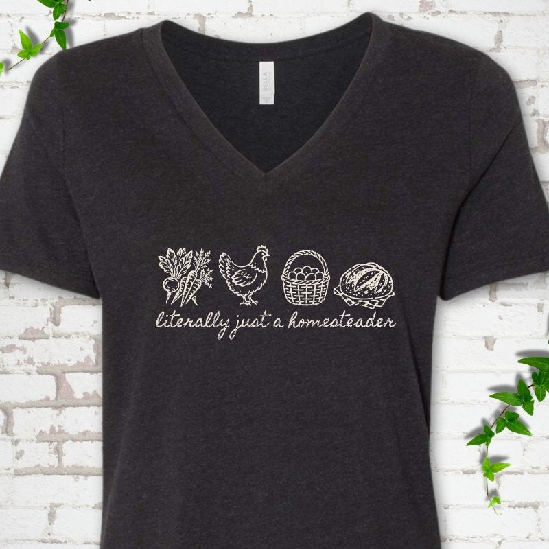Black Heather women's V-neck T-shirt flat lay with cream line-art icons in a row of carrots and radishes, a hen, a basket of eggs and a loaf of bread above the script words "literally just a homesteader", on white brick with ivy sprigs.