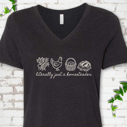 Black Heather women's V-neck T-shirt flat lay with cream line-art icons in a row of carrots and radishes, a hen, a basket of eggs and a loaf of bread above the script words "literally just a homesteader", on white brick with ivy sprigs.
