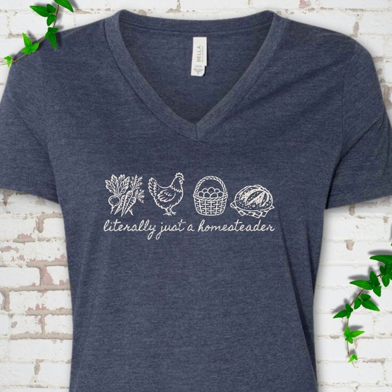 Heather Navy women's V-neck T-shirt flat lay with cream line-art icons in a row of carrots and radishes, a hen, a basket of eggs and a loaf of bread above the script words "literally just a homesteader", on white brick with ivy sprigs.