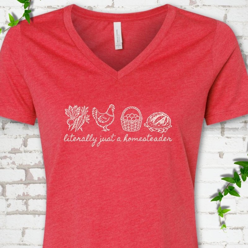 Heather Red women's V-neck T-shirt flat lay with white line-art icons in a row of carrots and radishes, a hen, a basket of eggs and a loaf of bread above the script words "literally just a homesteader", on white brick with ivy sprigs.