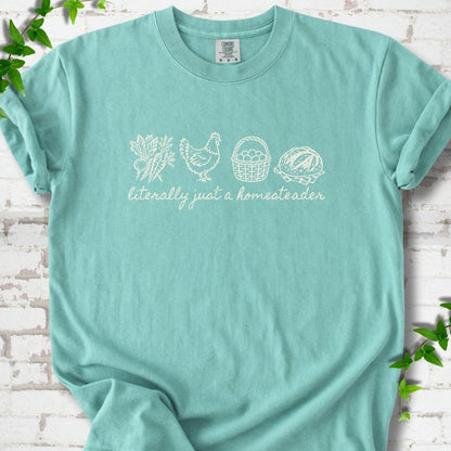 Chalky Mint T-shirt flat lay with cream line-art icons in a row of carrots and radishes, a hen, a basket of eggs and a loaf of bread above the script words "literally just a homesteader", on white brick with ivy sprigs.