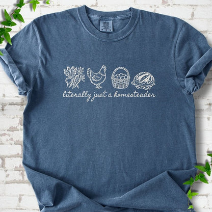 Denim T-shirt flat lay with cream line-art icons in a row of carrots and radishes, a hen, a basket of eggs and a loaf of bread above the script words "literally just a homesteader", on white brick with ivy sprigs.