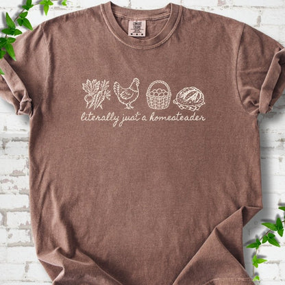Espresso T-shirt flat lay with cream line-art icons in a row of carrots and radishes, a hen, a basket of eggs and a loaf of bread above the script words "literally just a homesteader", on white brick with ivy sprigs.