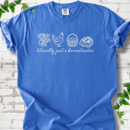 Flo Blue T-shirt flat lay with white line-art icons in a row of carrots and radishes, a hen, a basket of eggs and a loaf of bread above the script words "literally just a homesteader", on white brick with ivy sprigs.