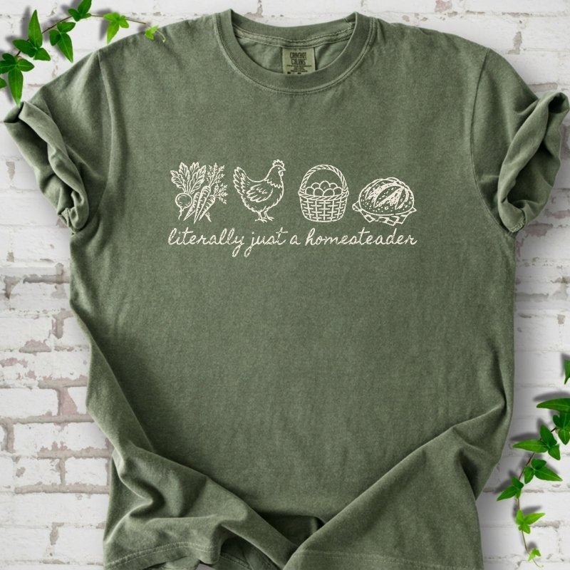 Moss T-shirt flat lay with cream line-art icons in a row of carrots and radishes, a hen, a basket of eggs and a loaf of bread above the script words "literally just a homesteader", on white brick with ivy sprigs.