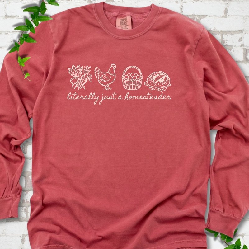 Crimson long sleeve T-shirt flat lay with cream line-art icons in a row of carrots and radishes, a hen, a basket of eggs and a loaf of bread above the script words "literally just a homesteader", on white brick with ivy sprigs.