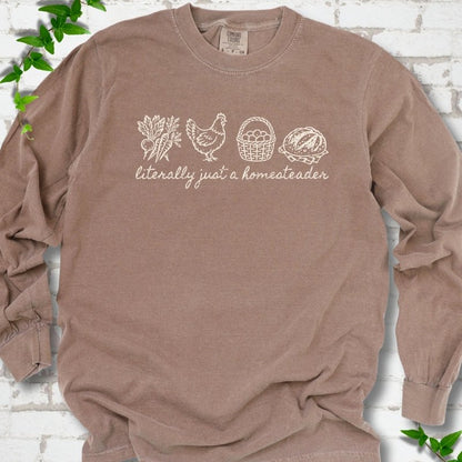 Espresso long sleeve T-shirt flat lay with cream line-art icons in a row of carrots and radishes, a hen, a basket of eggs and a loaf of bread above the script words "literally just a homesteader", on white brick with ivy sprigs.