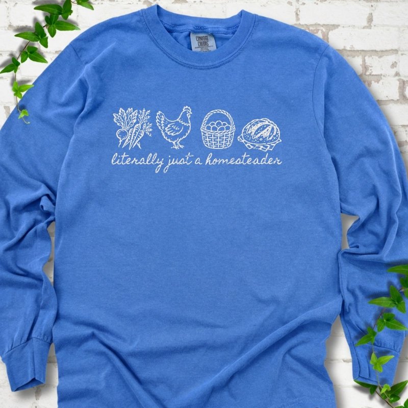 Flo Blue long sleeve T-shirt flat lay with white line-art icons in a row of carrots and radishes, a hen, a basket of eggs and a loaf of bread above the script words "literally just a homesteader", on white brick with ivy sprigs.
