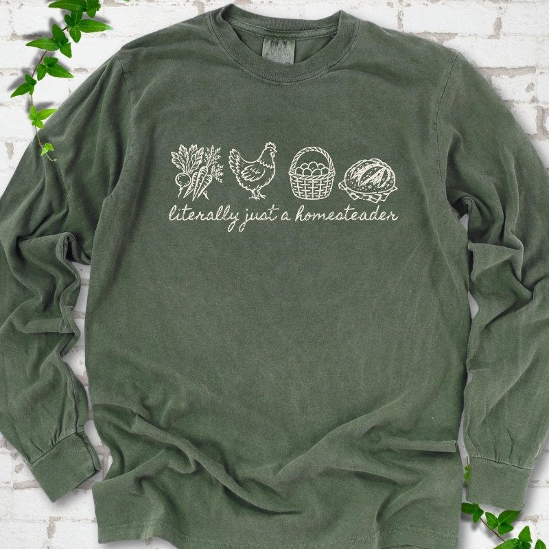 Hemp long sleeve T-shirt flat lay with cream line-art icons in a row of carrots and radishes, a hen, a basket of eggs and a loaf of bread above the script words "literally just a homesteader", on white brick with ivy sprigs.