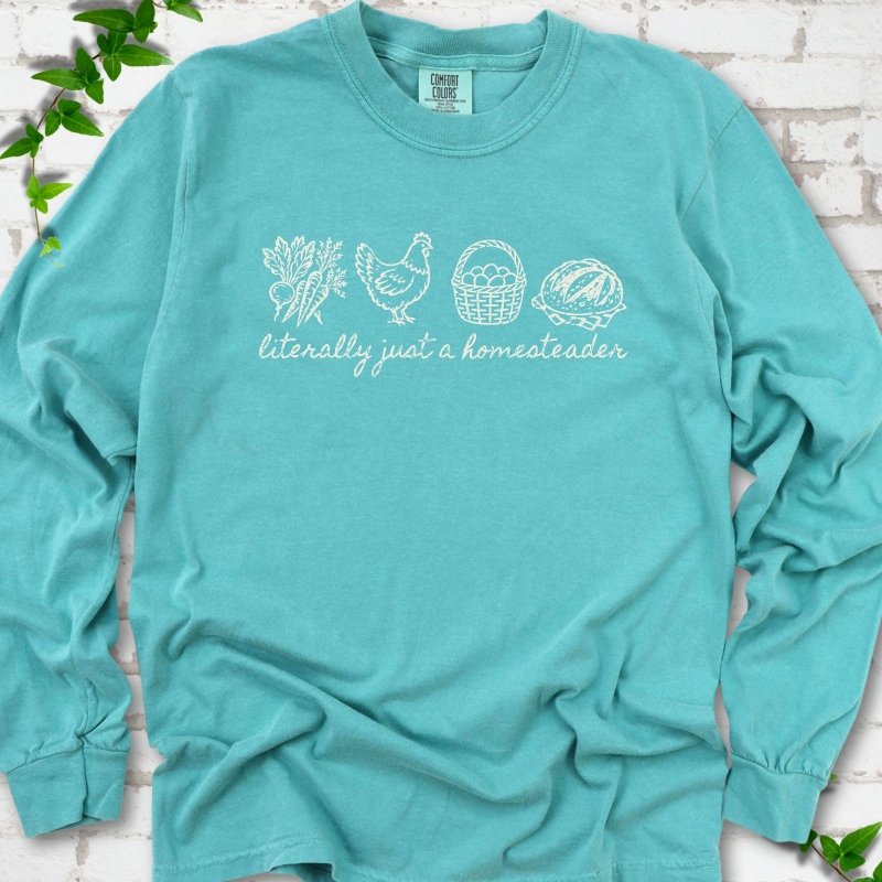Seafoam long sleeve T-shirt flat lay with white line-art icons in a row of carrots and radishes, a hen, a basket of eggs and a loaf of bread above the script words "literally just a homesteader", on white brick with ivy sprigs.
