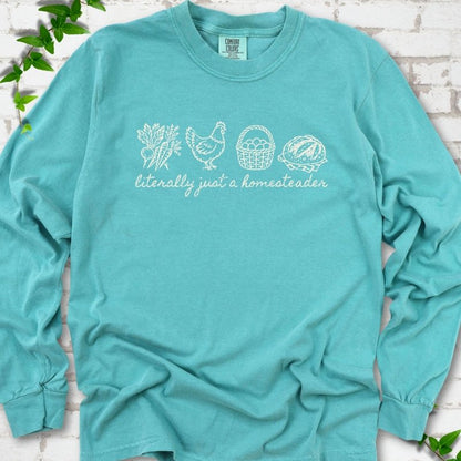 Seafoam long sleeve T-shirt flat lay with white line-art icons in a row of carrots and radishes, a hen, a basket of eggs and a loaf of bread above the script words "literally just a homesteader", on white brick with ivy sprigs.