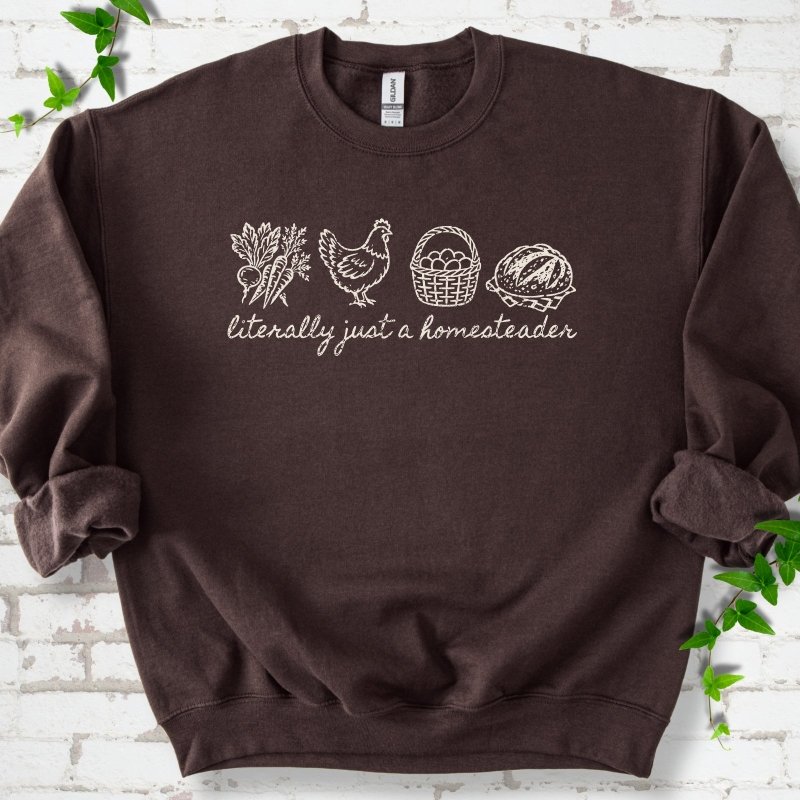 Dark Chocolate sweatshirt flat lay with cream line-art icons in a row of carrots and radishes, a hen, a basket of eggs and a loaf of bread above the script words "literally just a homesteader", on white brick with ivy sprigs.