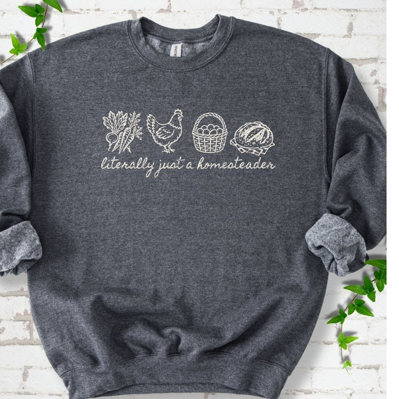 Dark Heather sweatshirt flat lay with cream line-art icons in a row of carrots and radishes, a hen, a basket of eggs and a loaf of bread above the script words "literally just a homesteader", on white brick with ivy sprigs.