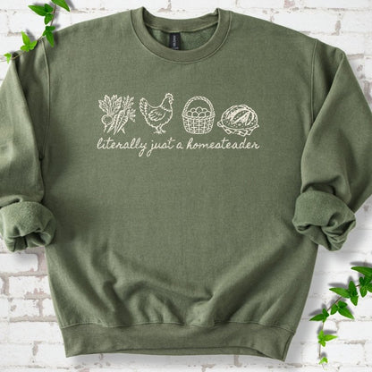 Military Green sweatshirt flat lay with cream line-art icons in a row of carrots and radishes, a hen, a basket of eggs and a loaf of bread above the script words "literally just a homesteader", on white brick with ivy sprigs.