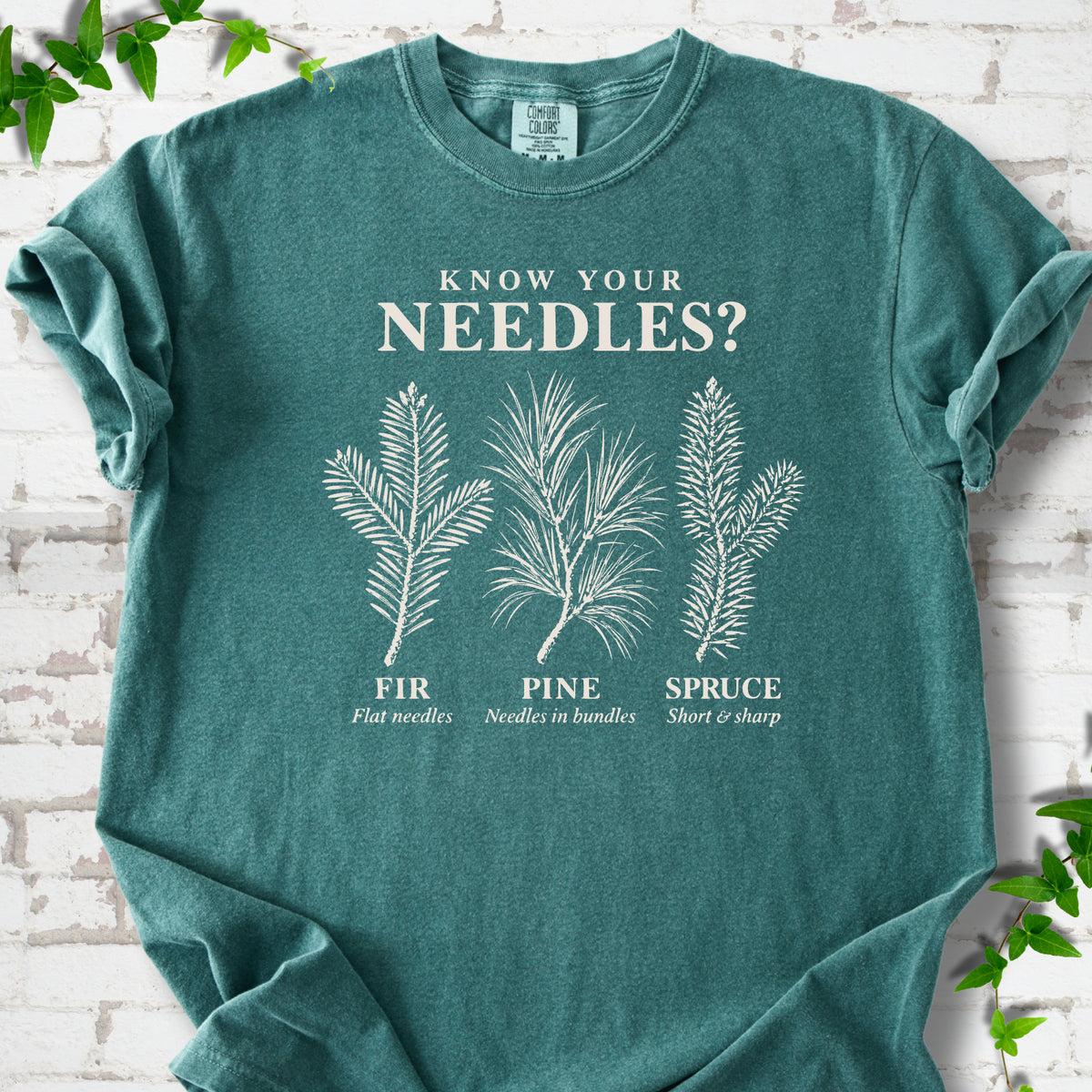Blue Spruce T-shirt laid flat, showing cream fir, pine and spruce needle illustrations beneath "Know Your Needles?" with identification labels.