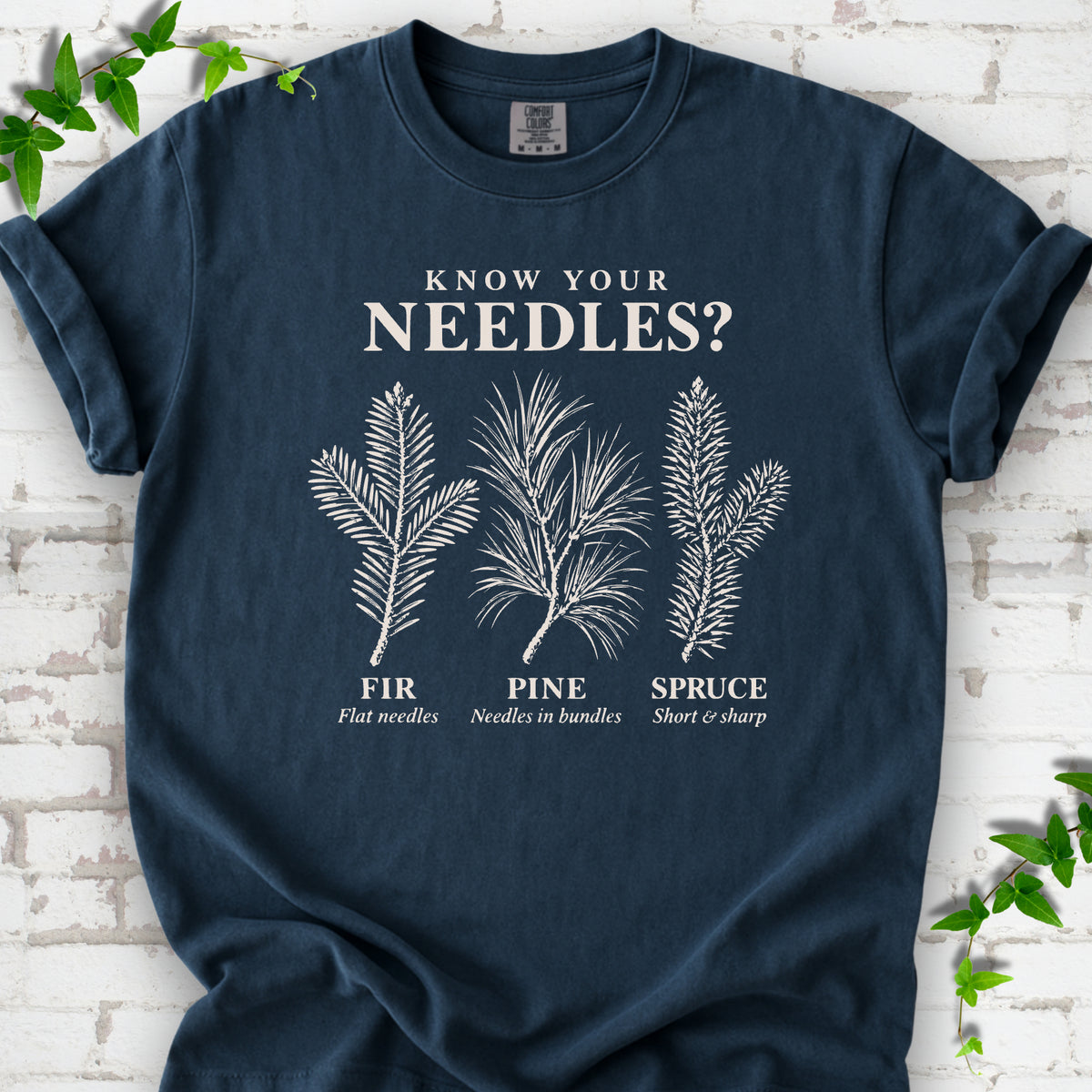 Midnight T-shirt laid flat, showing cream fir, pine and spruce needle illustrations beneath "Know Your Needles?" with identification labels.