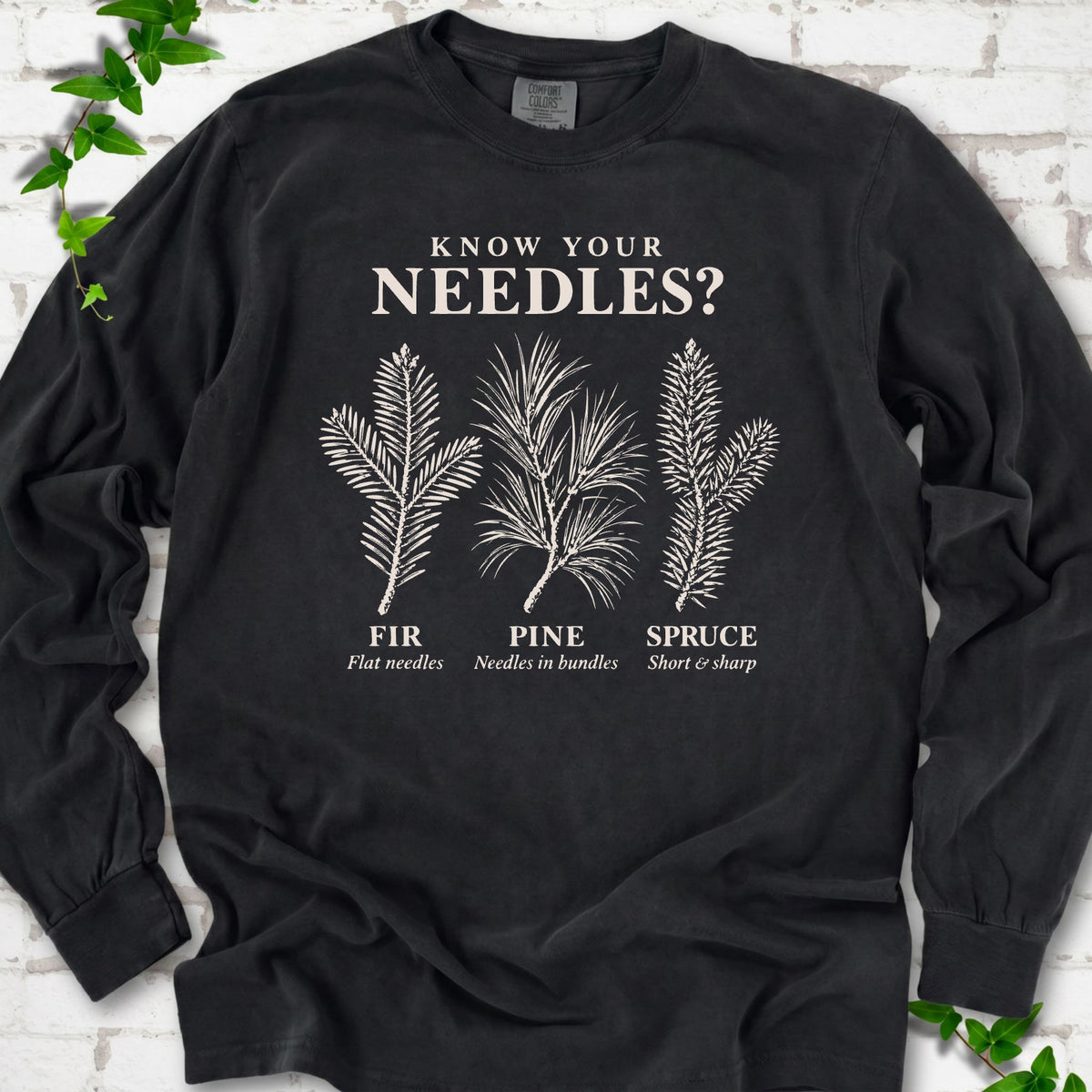 Black long sleeve tee laid flat, showing cream fir, pine and spruce needle illustrations beneath "Know Your Needles?" with identification labels.