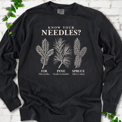 Black long sleeve tee laid flat, showing cream fir, pine and spruce needle illustrations beneath "Know Your Needles?" with identification labels.