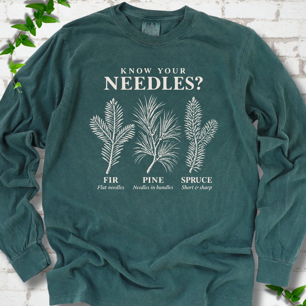 Blue Spruce long sleeve tee laid flat, showing cream fir, pine and spruce needle illustrations beneath "Know Your Needles?" with identification labels.