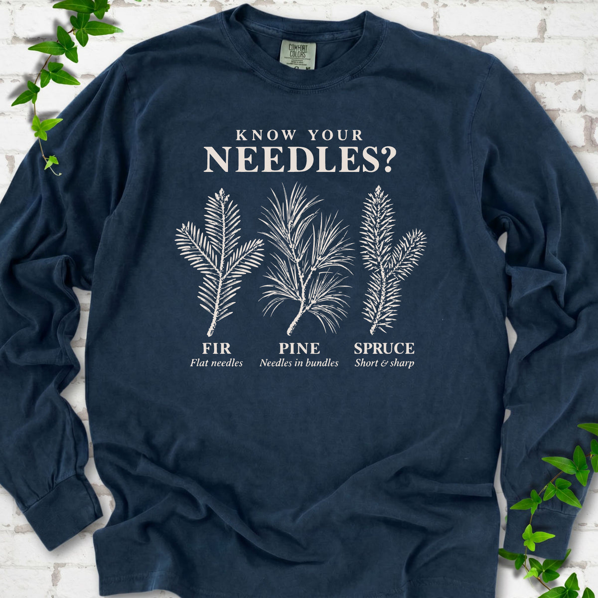Midnight long sleeve tee laid flat, showing cream fir, pine and spruce needle illustrations beneath "Know Your Needles?" with identification labels.