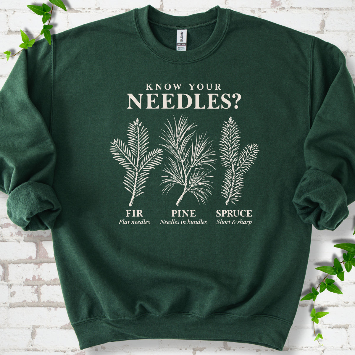 Forest Green classic sweatshirt laid flat, showing cream fir, pine and spruce needle illustrations beneath "Know Your Needles?" with identification labels.