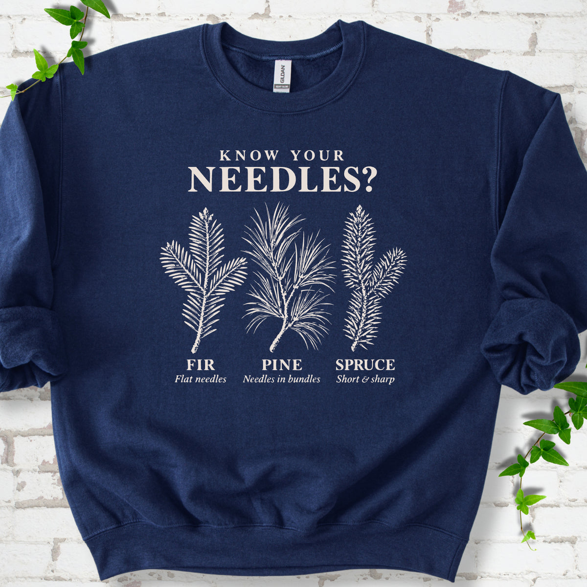 Navy classic sweatshirt laid flat, showing cream fir, pine and spruce needle illustrations beneath "Know Your Needles?" with identification labels.