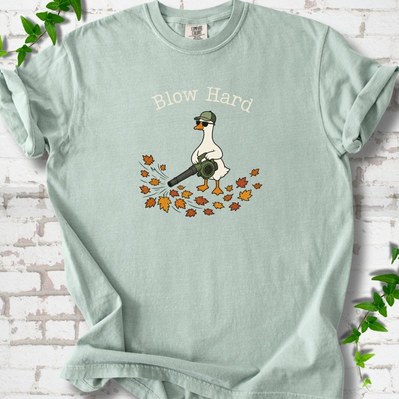 Bay T-shirt flat lay with ivory "Blow Hard" lettering above a white goose in a green cap and sunglasses using a leaf blower on a pile of orange and gold fall leaves, on white brick with ivy sprigs.