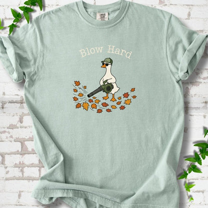 Bay T-shirt flat lay with ivory "Blow Hard" lettering above a white goose in a green cap and sunglasses using a leaf blower on a pile of orange and gold fall leaves, on white brick with ivy sprigs.