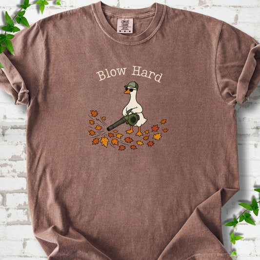 Espresso T-shirt flat lay with ivory "Blow Hard" lettering above a white goose in a green cap and sunglasses using a leaf blower on a pile of orange and gold fall leaves, on white brick with ivy sprigs.