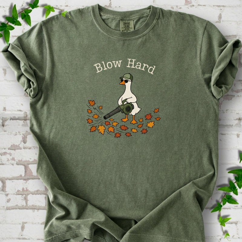 Moss T-shirt flat lay with ivory "Blow Hard" lettering above a white goose in a green cap and sunglasses using a leaf blower on a pile of orange and gold fall leaves, on white brick with ivy sprigs.