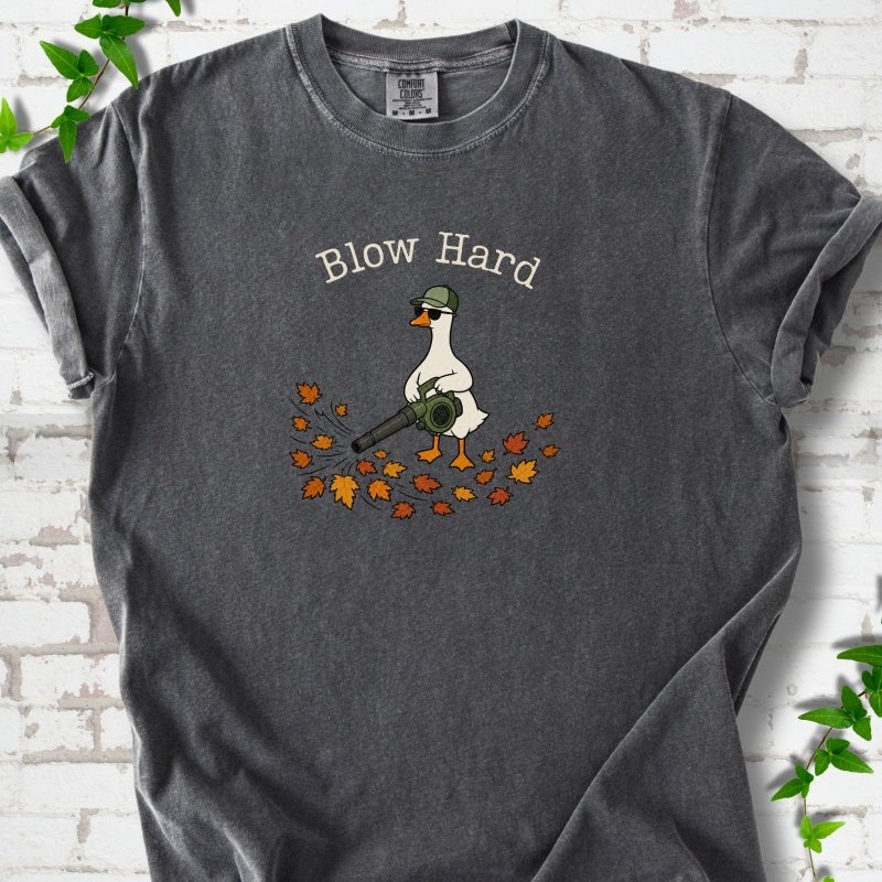 Pepper T-shirt flat lay with ivory "Blow Hard" lettering above a white goose in a green cap and sunglasses using a leaf blower on a pile of orange and gold fall leaves, on white brick with ivy sprigs.