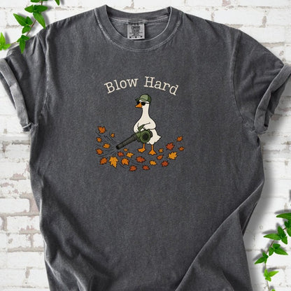Pepper T-shirt flat lay with ivory "Blow Hard" lettering above a white goose in a green cap and sunglasses using a leaf blower on a pile of orange and gold fall leaves, on white brick with ivy sprigs.