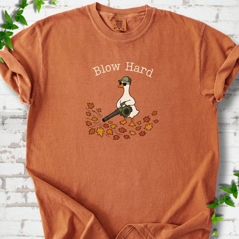 Yam T-shirt flat lay with ivory "Blow Hard" lettering above a white goose in a green cap and sunglasses using a leaf blower on a pile of orange and gold fall leaves, on white brick with ivy sprigs.
